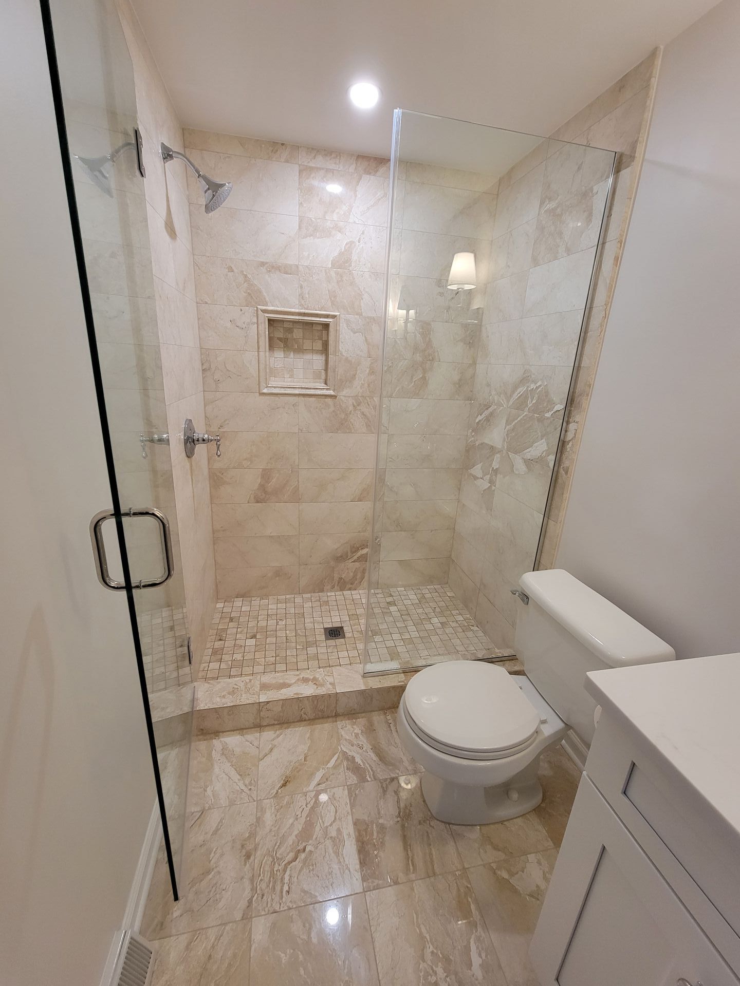 Bathroom with tiled shower, glass door, toilet, and vanity. Beige and light brown tones dominate.