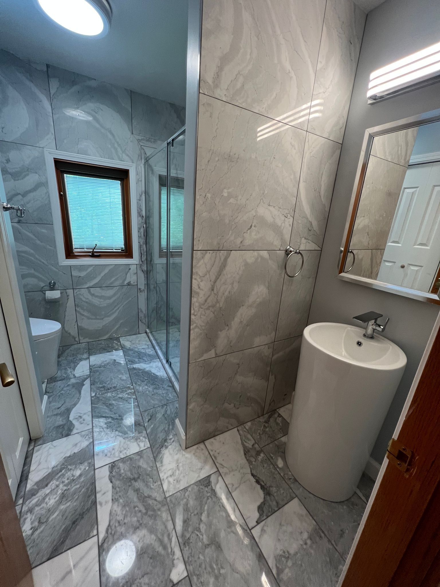 Modern bathroom with gray marble-look tile, glass shower, pedestal sink, toilet, and window.