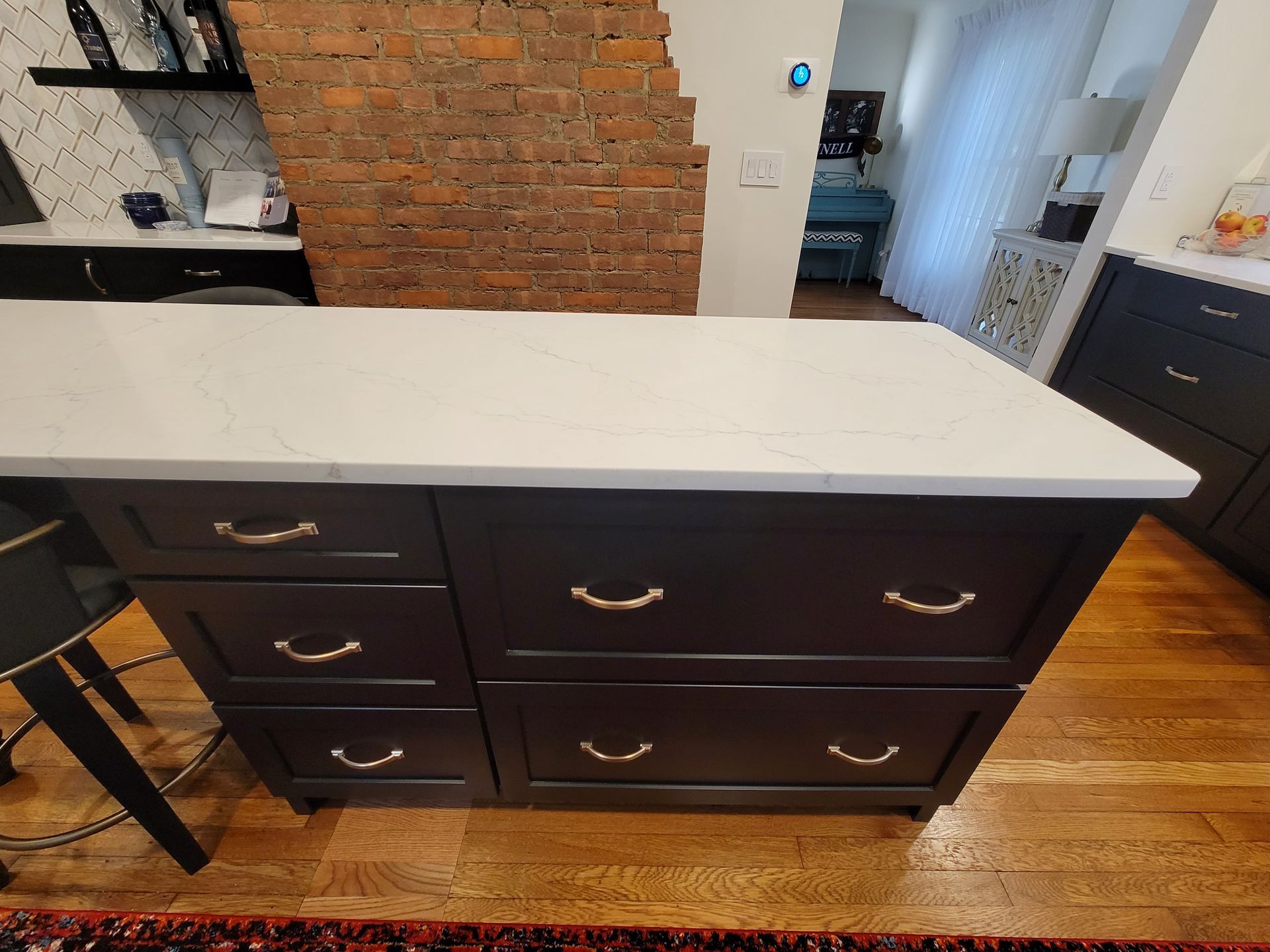 Kitchen island with dark cabinets, gold handles, and white countertop. Exposed brick wall and wood floor in the background.