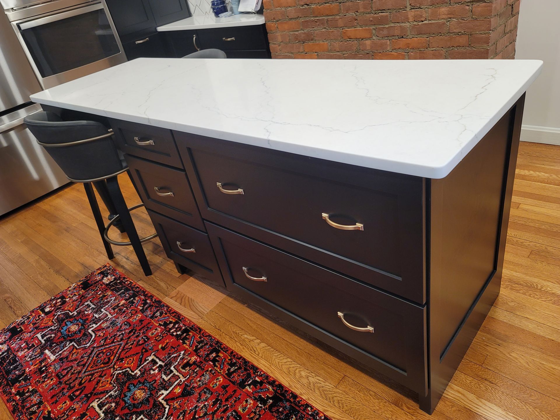 Kitchen island with white countertop, dark cabinets, and brass handles. A red rug and stool are nearby.