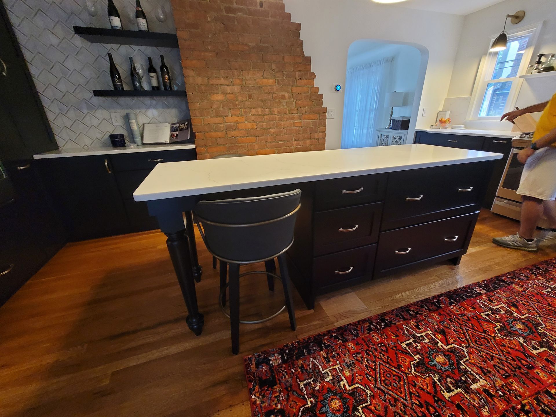 Kitchen island with drawers, white countertop, dark cabinets, a stool, and a red rug.