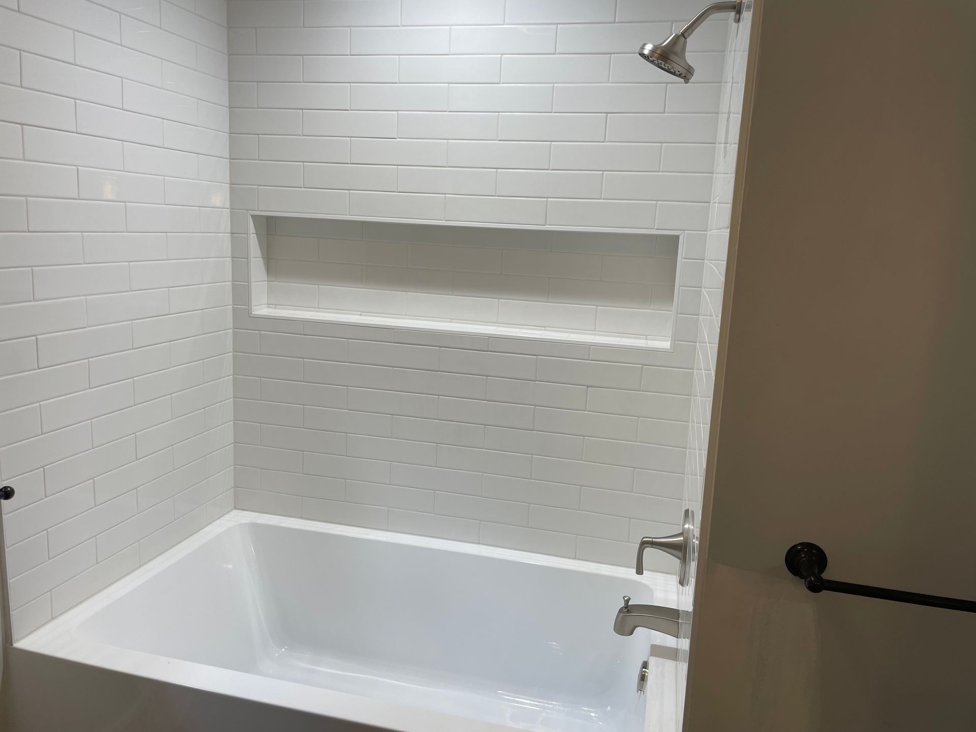 White-tiled bathroom with a built-in shelf, showerhead, and bathtub, against a tan wall.