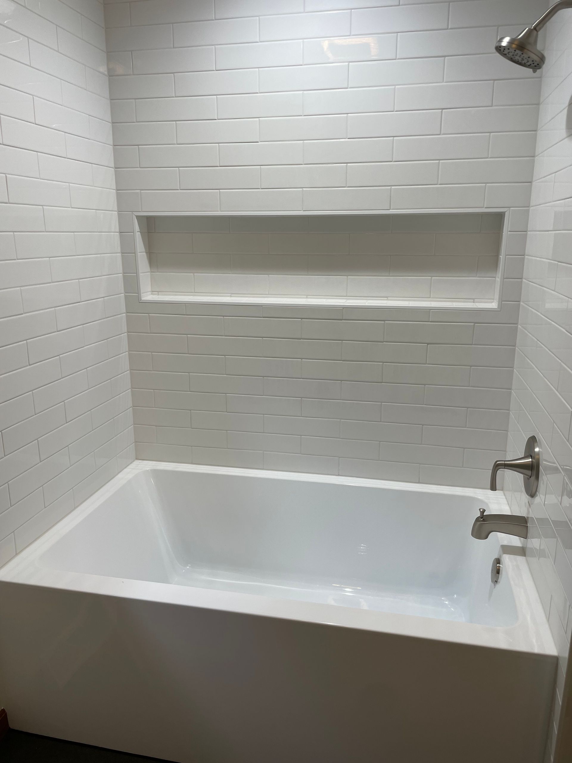 White tiled shower with built-in shelf above a white bathtub, silver showerhead and faucet.