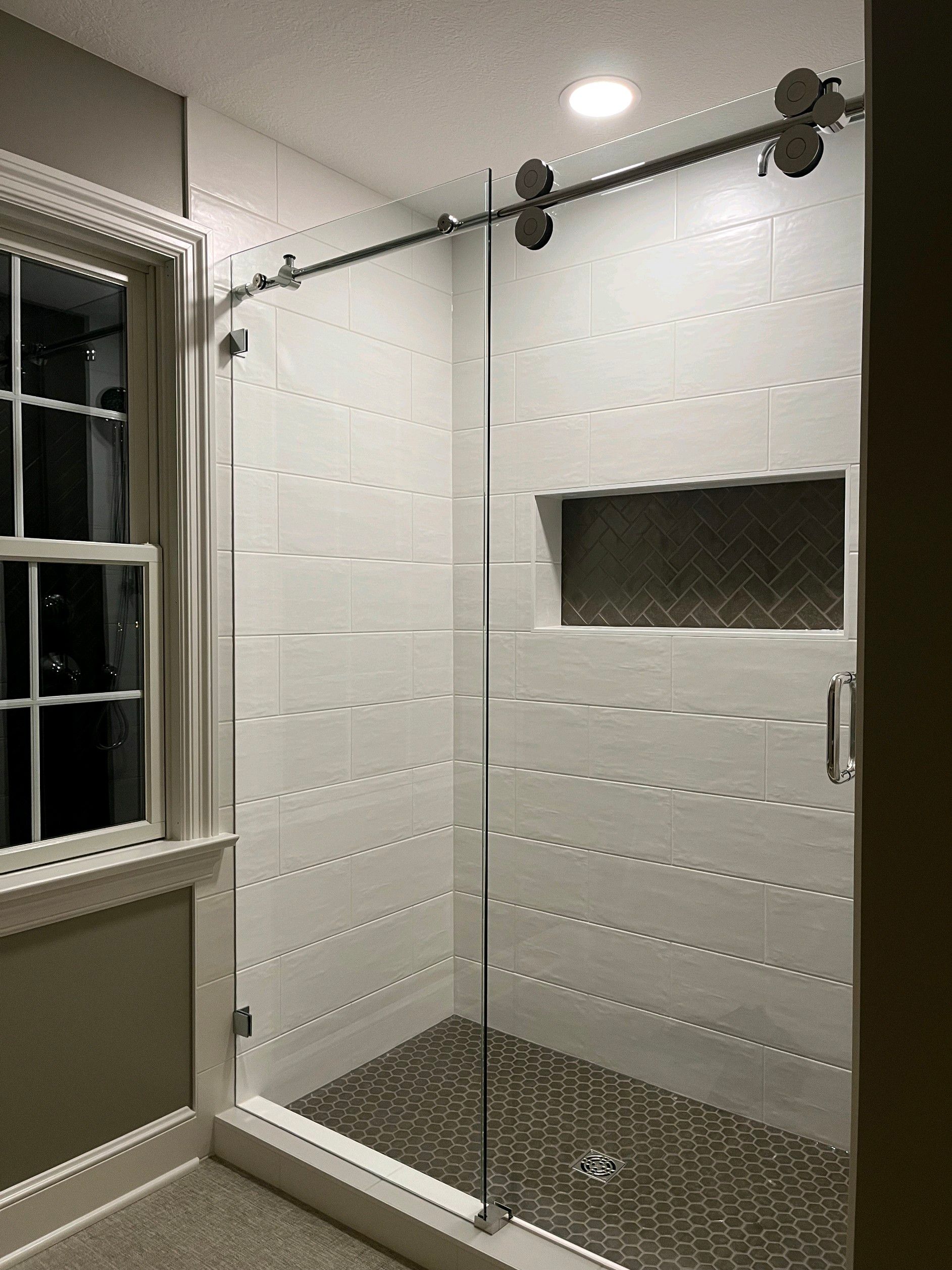 Modern bathroom with glass shower enclosure, white tiled walls, dark floor, window on the left.