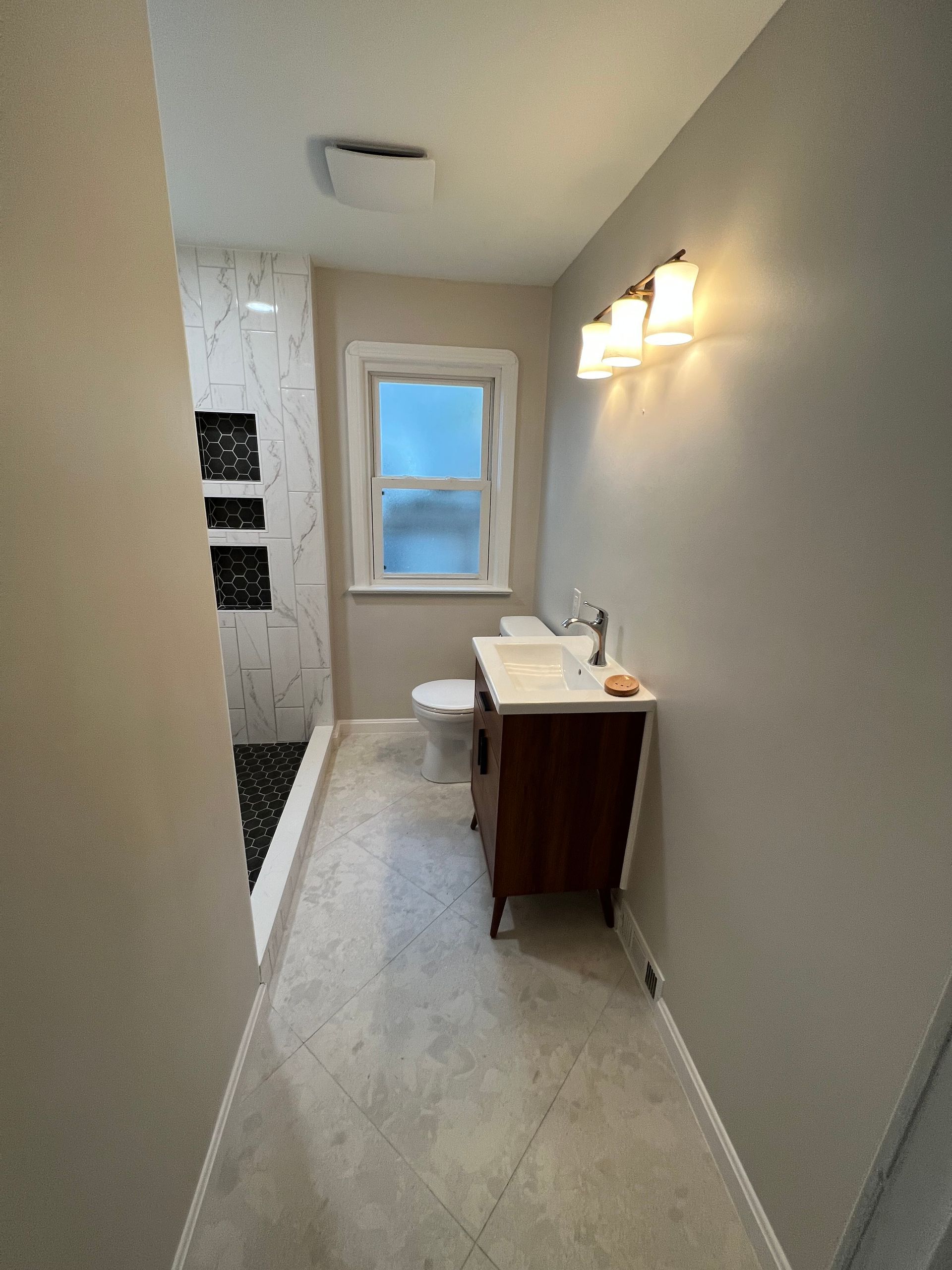 Bathroom with gray walls, white sink, wooden cabinet, and window. Shower with black and white tile.