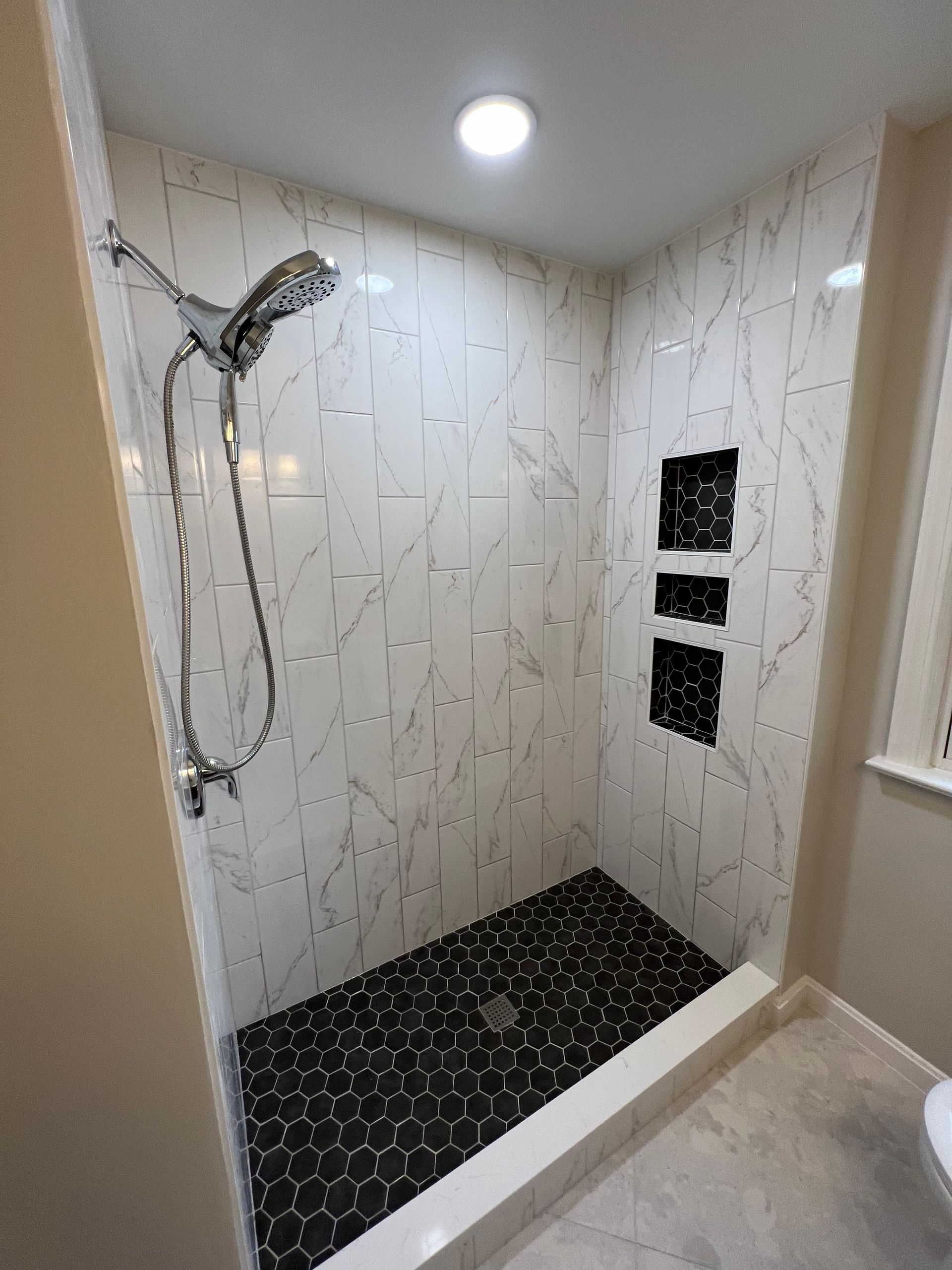 A tiled shower with black pebble floor and white marble-look walls, chrome showerhead, and inset shelves.