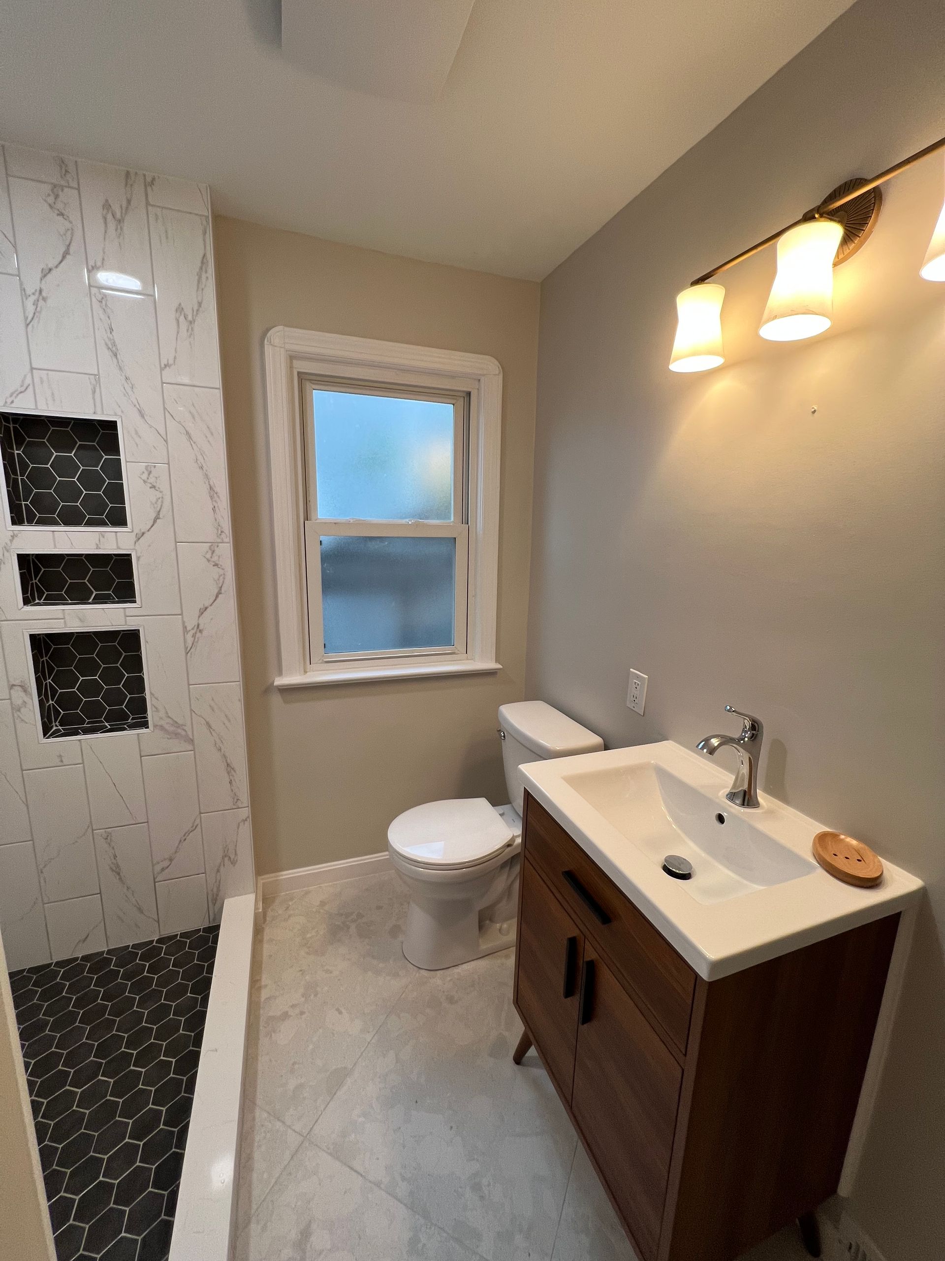 Bathroom with shower, toilet, and vanity. Gray walls, light fixture, window. Shower has marble and black tile.