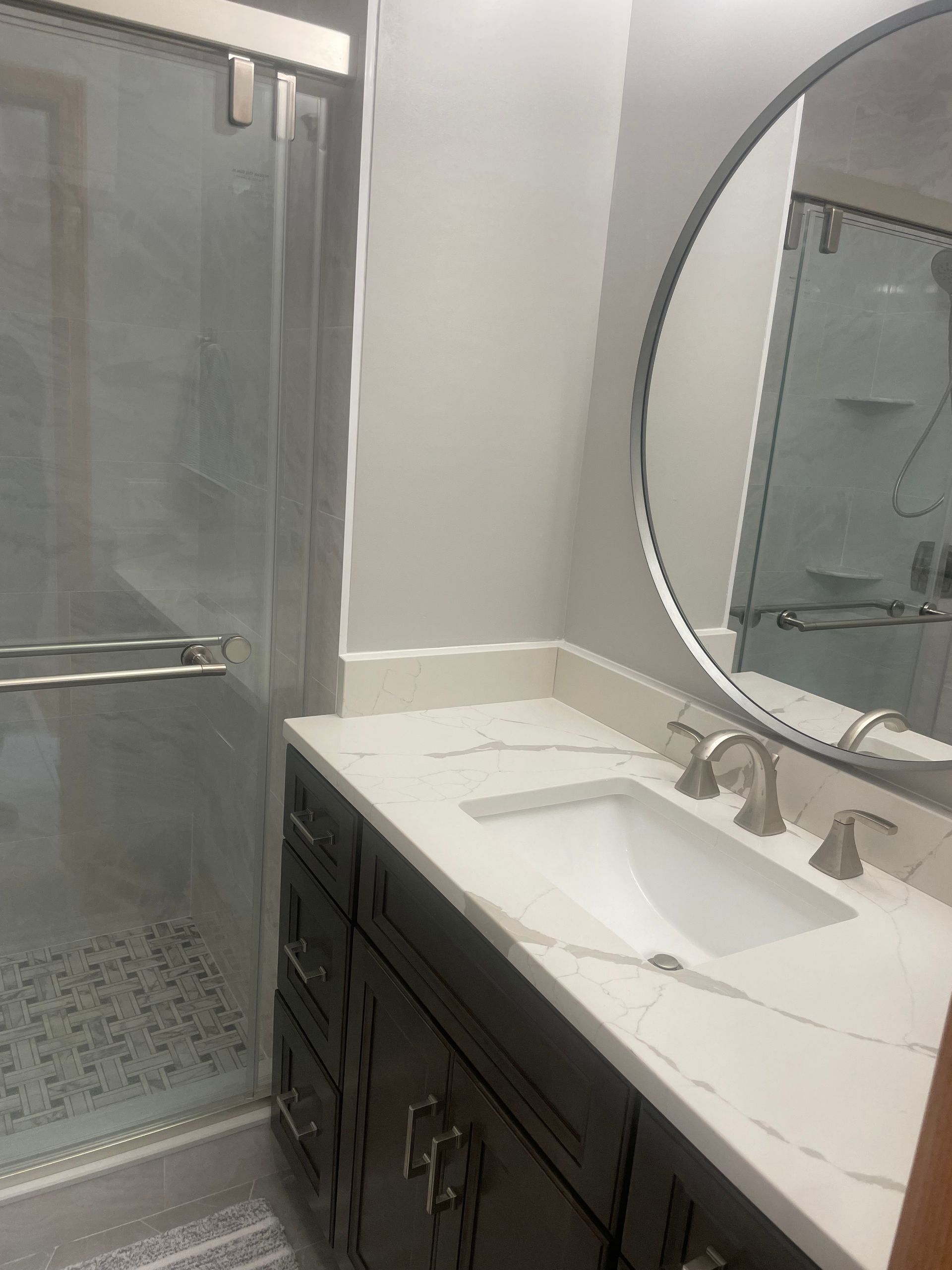 Bathroom with a dark vanity, white countertop, round mirror, and glass shower.
