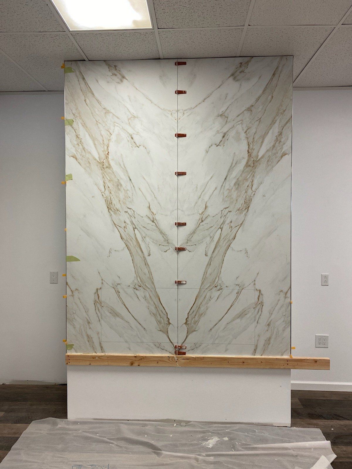 Large marble slab with symmetrical veining, displayed on a wall. Copper-colored clamps and a wooden ledge are present.