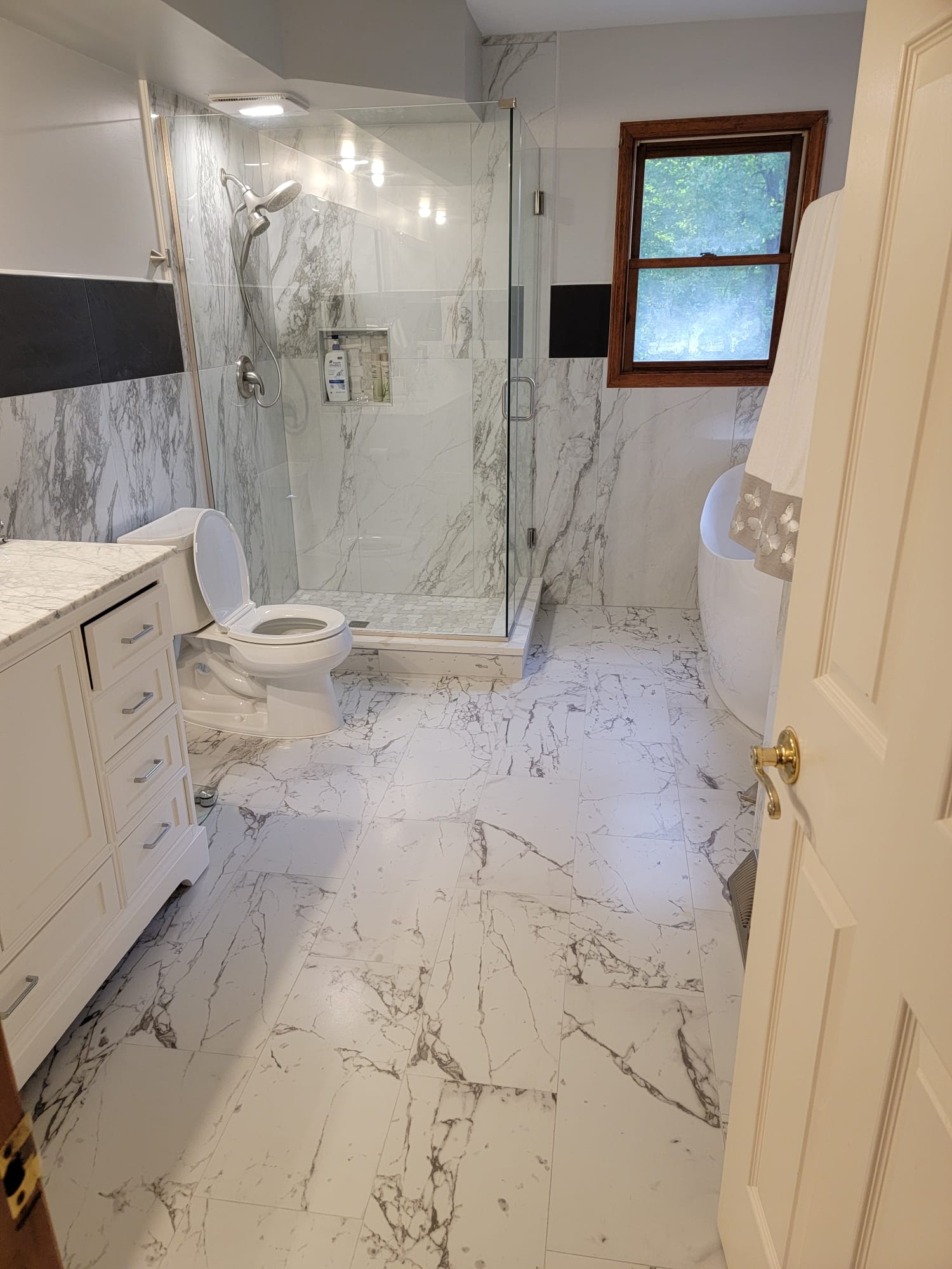Bathroom with marble tile, glass shower, white vanity, and tub. Brown-framed window.
