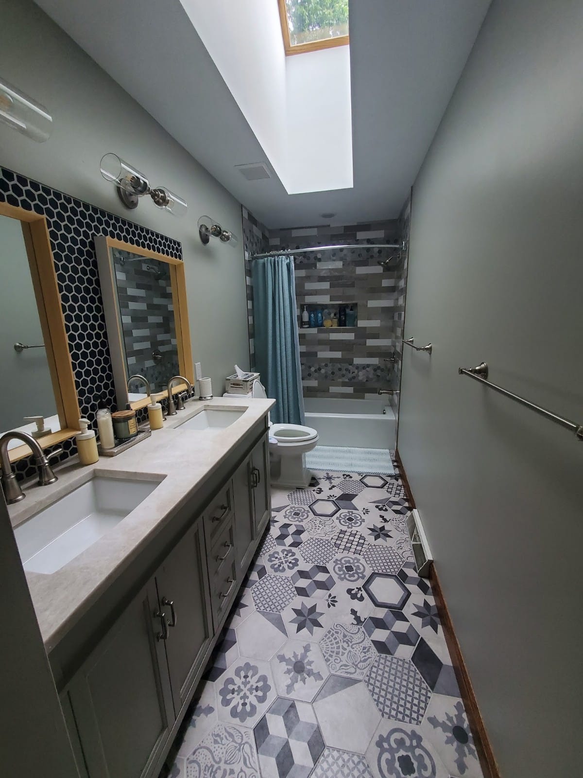Bathroom with double sinks, patterned floor, and shower. Gray, white, and blue tones. Skylight overhead.