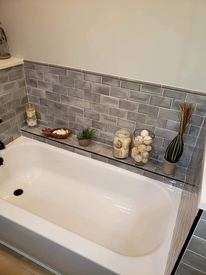 Bathroom with a white bathtub, gray tiled wall, and glass shelf holding decorative items.