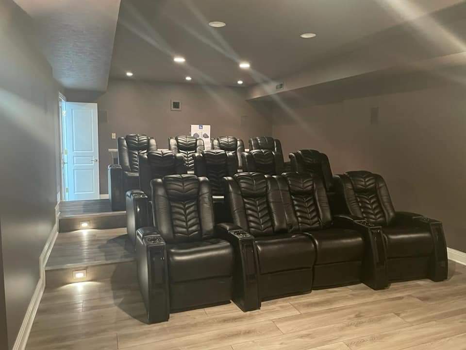 Home theater with tiered seating of black reclining chairs. Brown walls, recessed lighting, and light wood-look floor.