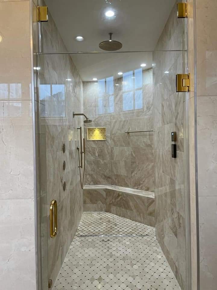 Spacious shower with glass door and gold hardware. Beige tile walls, bench, and mosaic tile floor.