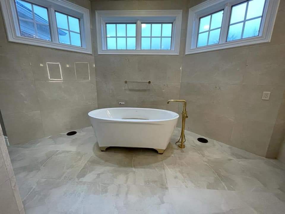 Bathroom with white, claw-footed tub, gold faucet, and large windows. Beige tile covers the walls and floor.