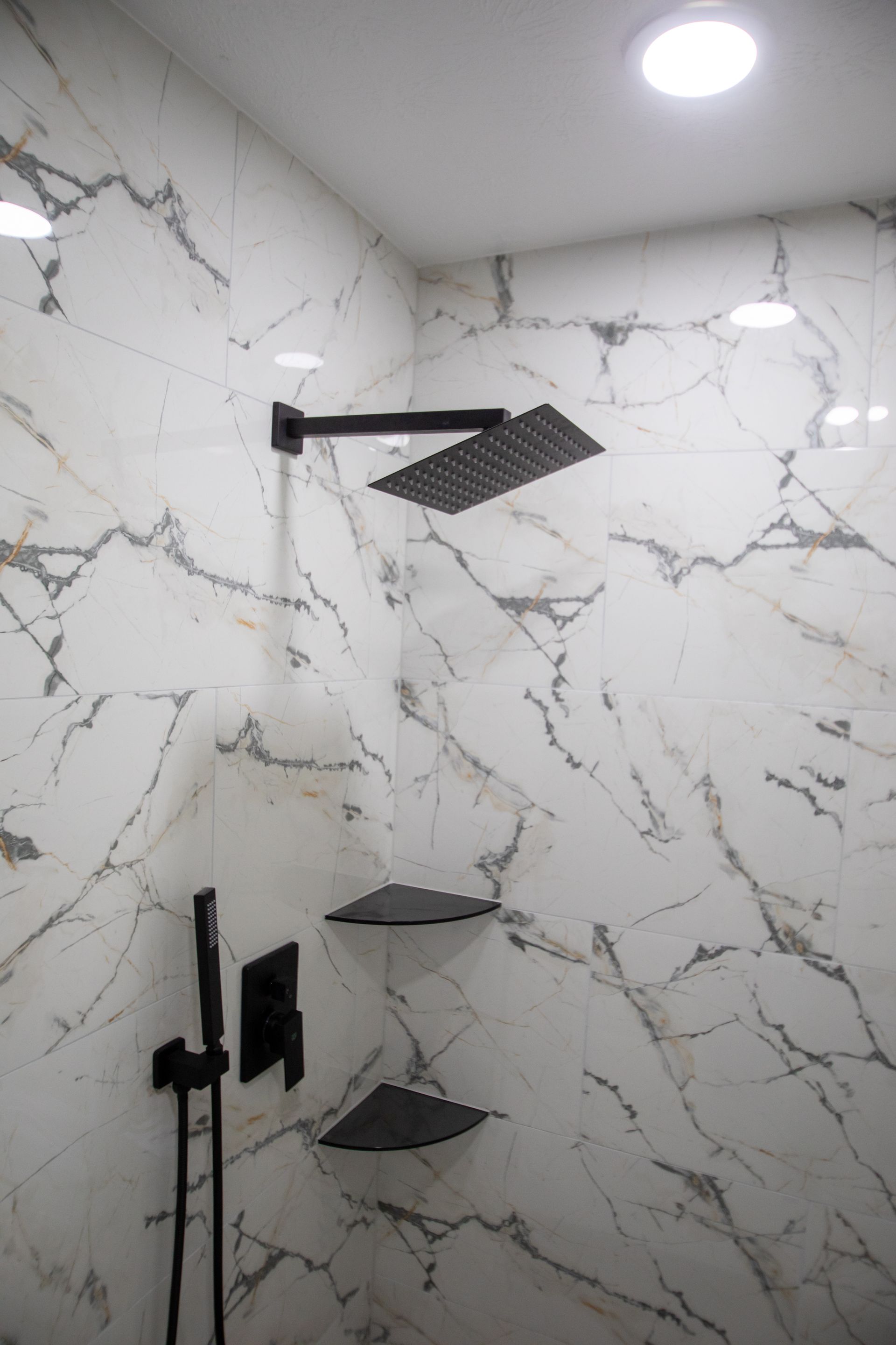 Black shower fixtures on marble-patterned tile wall with recessed ceiling lights and corner shelves.