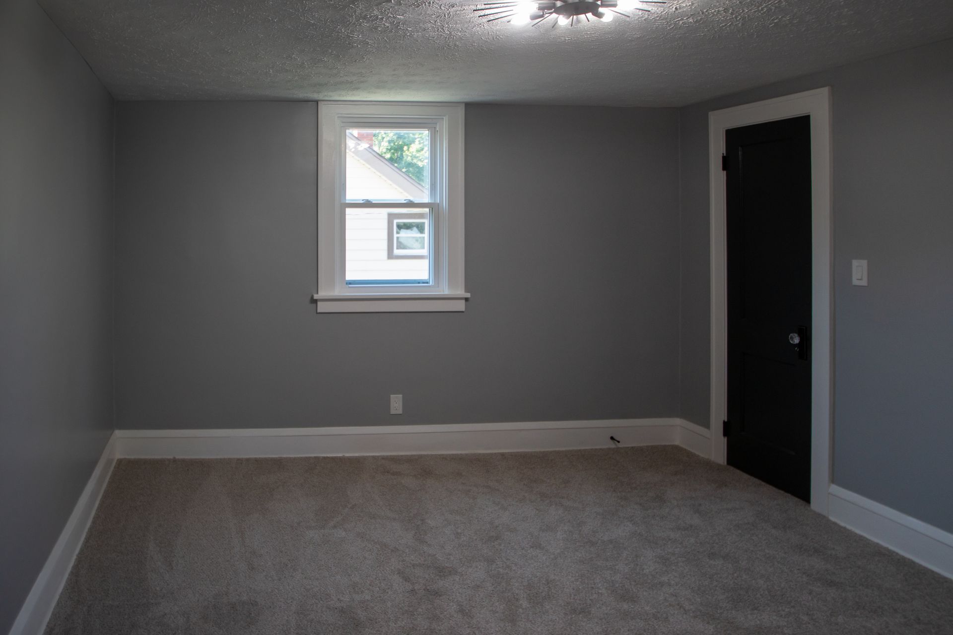 Empty room with gray walls, window, black door, white trim, and carpeted floor.