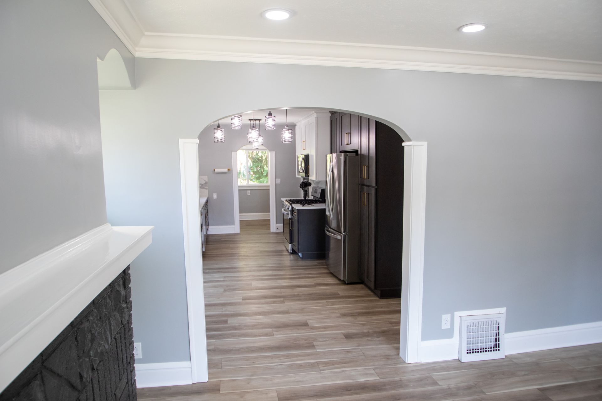 Light gray-walled interior with arched doorways leading to kitchen, wood floors, white trim, and a fireplace.