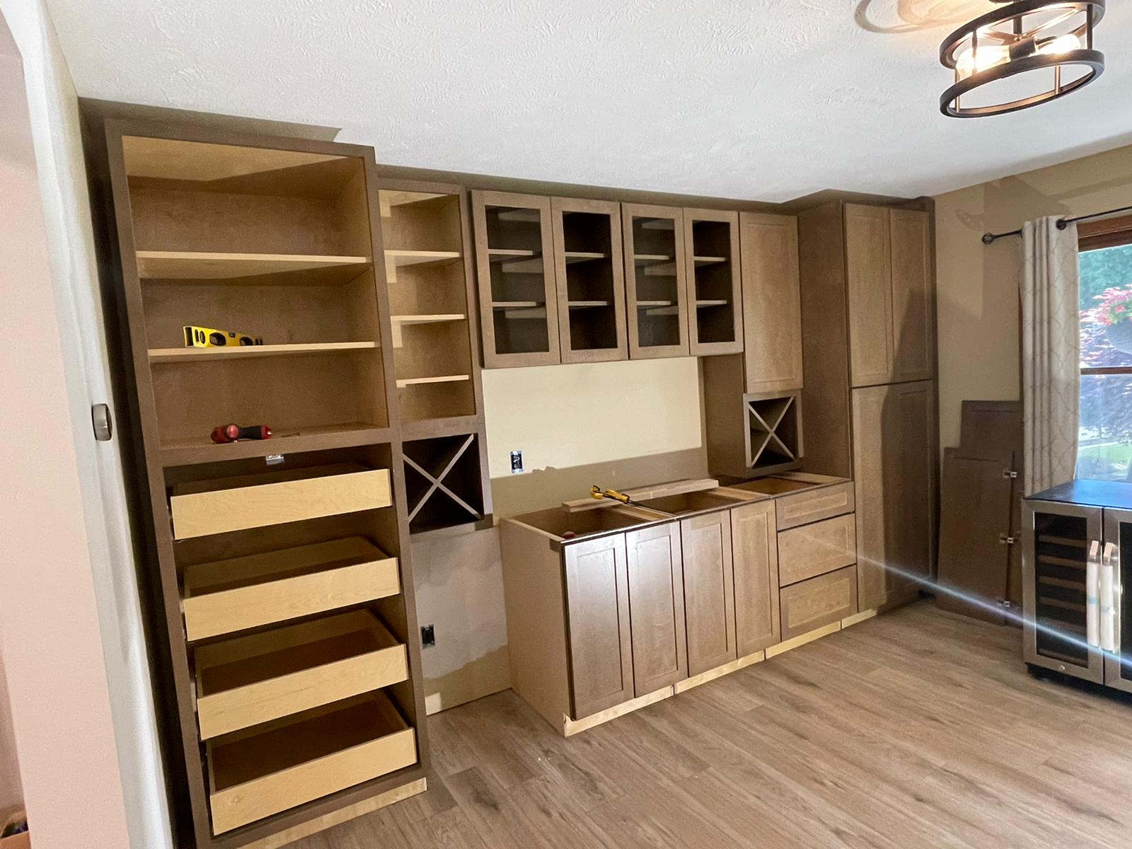 Kitchen cabinets under construction: brown wood, some with glass doors, drawers, shelves, wine rack, light wood floors.