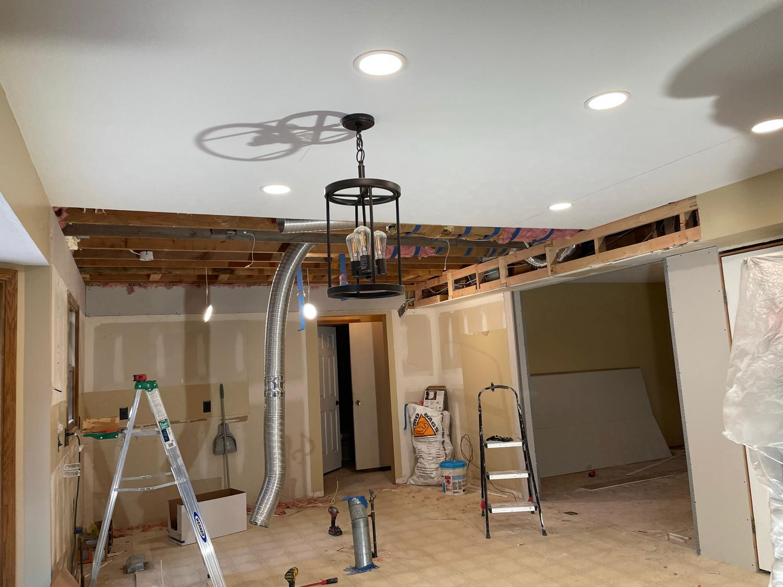 Room undergoing renovation: exposed ceiling beams, new recessed lighting, hanging light fixture. Drywall partially installed.