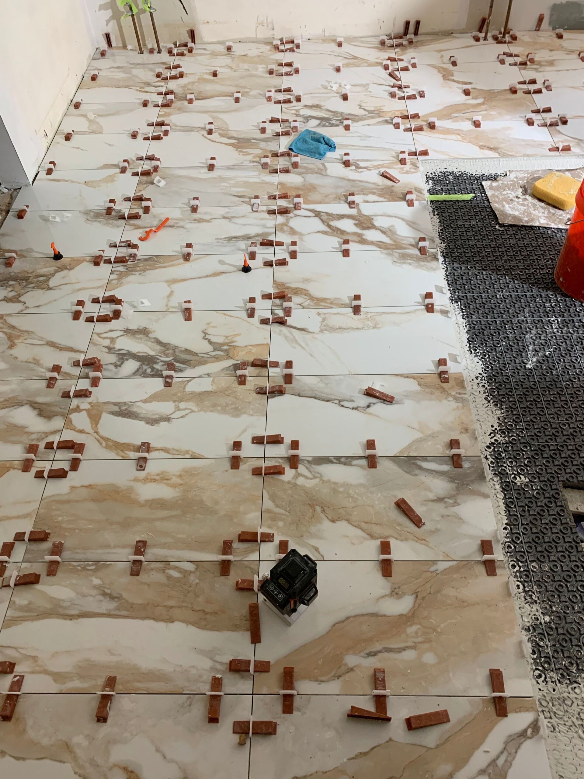 Floor tiles being installed, with leveling clips in place. White marble pattern, brown and tan accents. Construction site.