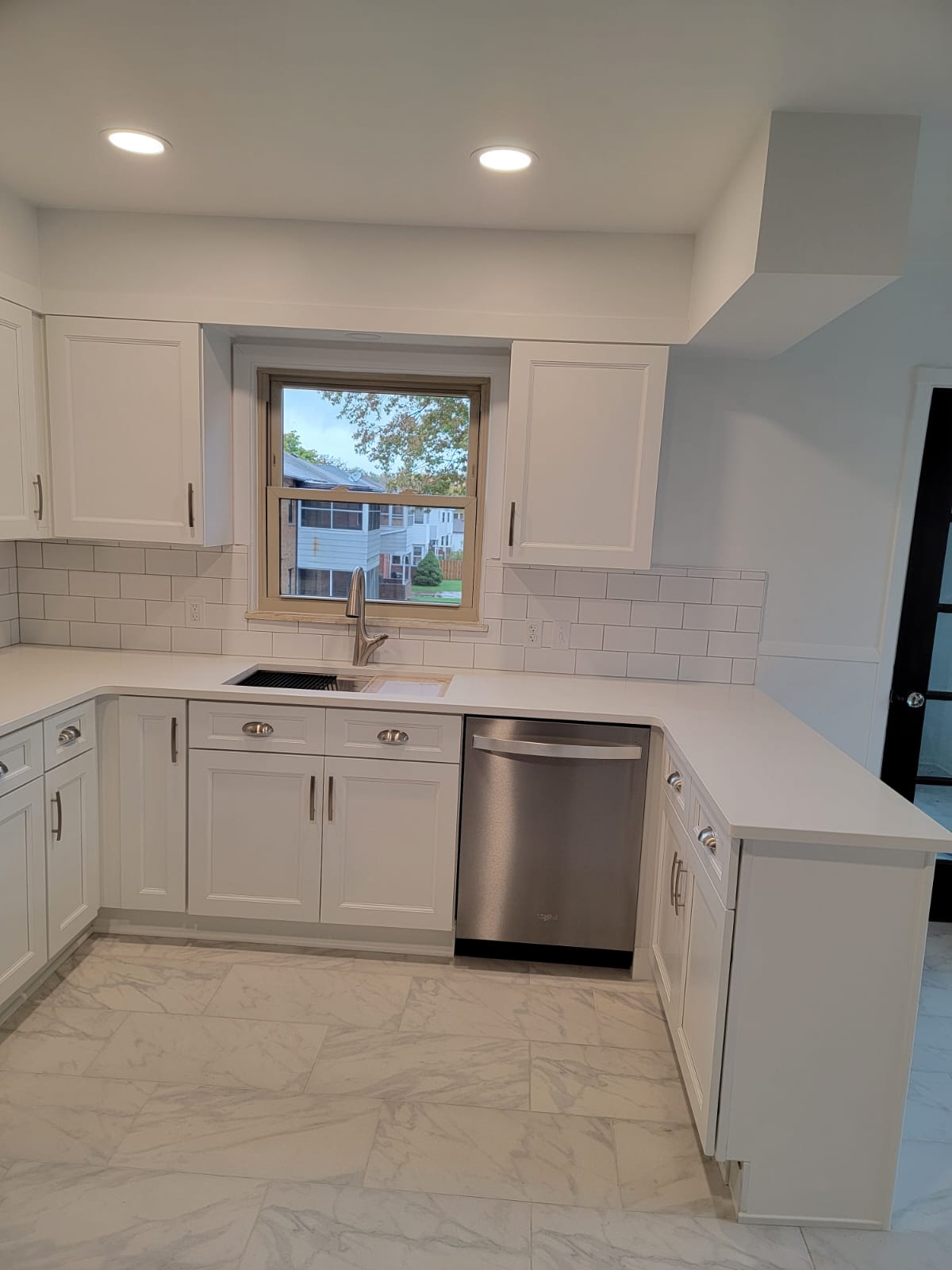 White kitchen with cabinets, countertops, and stainless steel dishwasher. Tile backsplash and window.