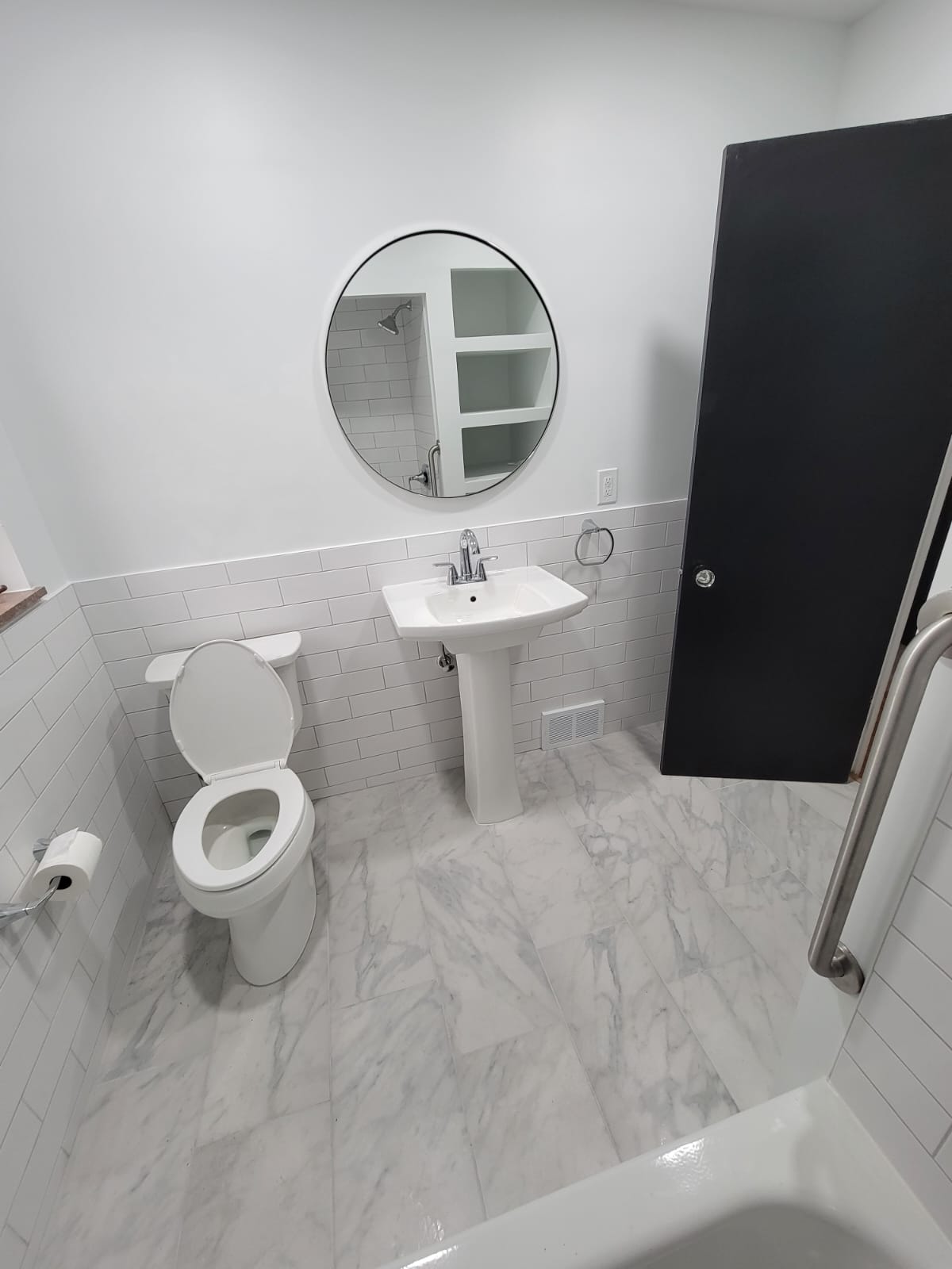 Bathroom interior: white walls, marble-look floor, pedestal sink, toilet, round mirror, black door, and a grab bar.