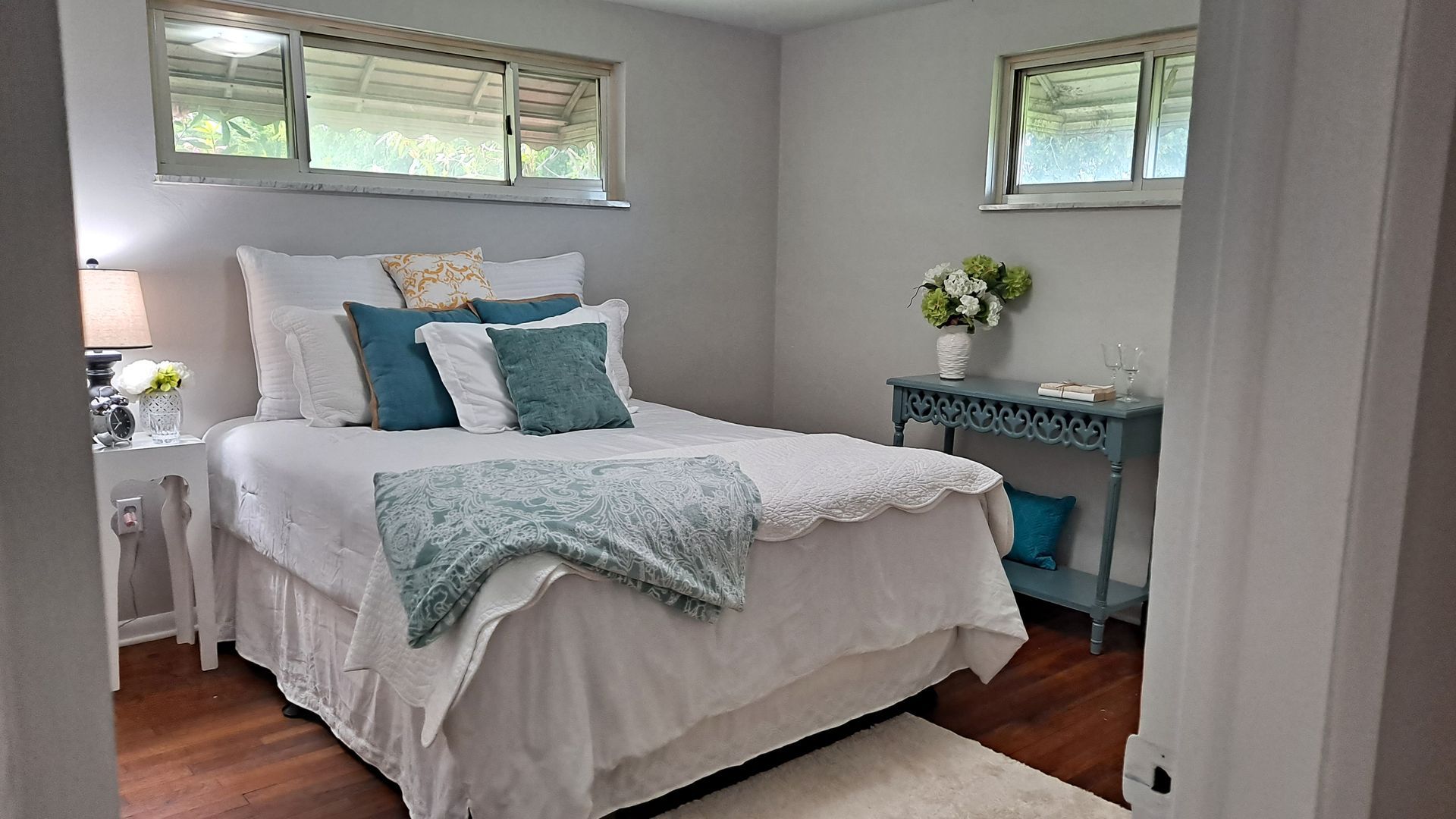 Bedroom with white bedding, turquoise pillows and blanket, and a small blue table with flowers.