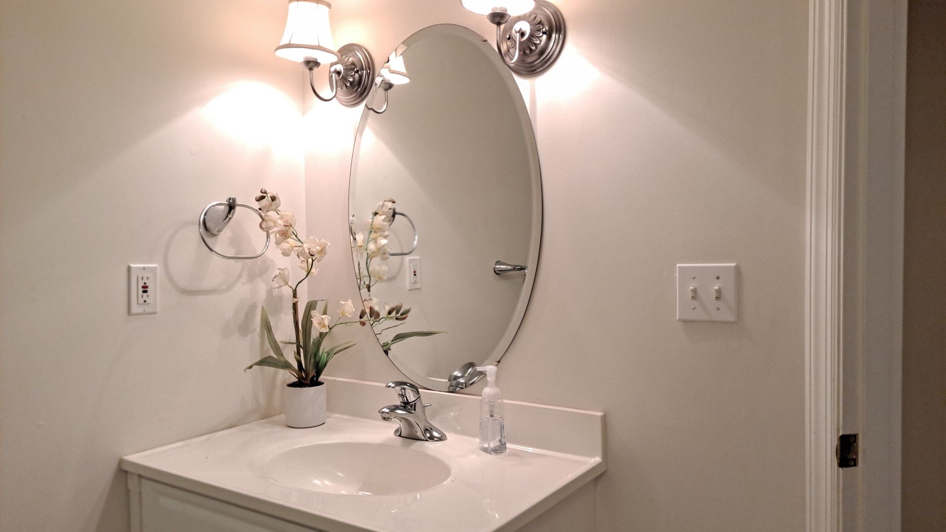 Bathroom with oval mirror, white sink, lights, and flower decor.