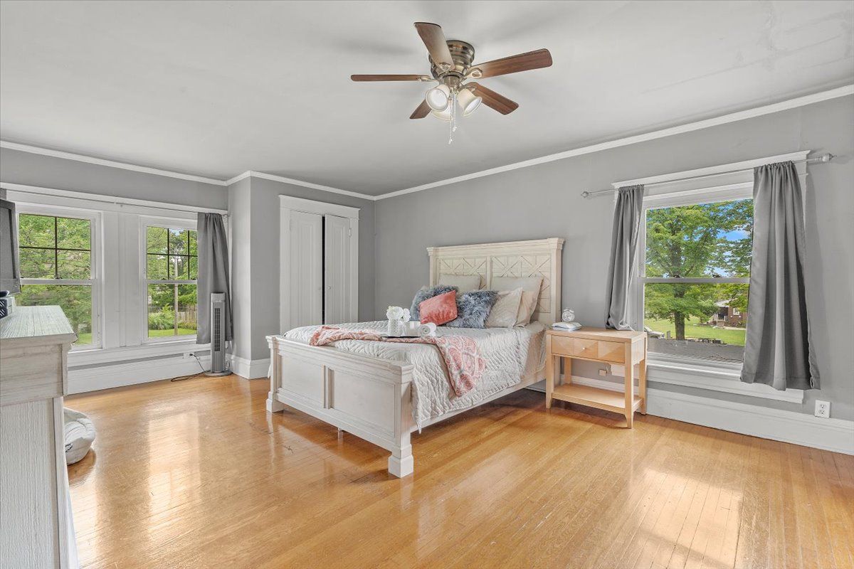 Bedroom with hardwood floors, light gray walls, white bed and trim, two windows with gray curtains, and a ceiling fan.