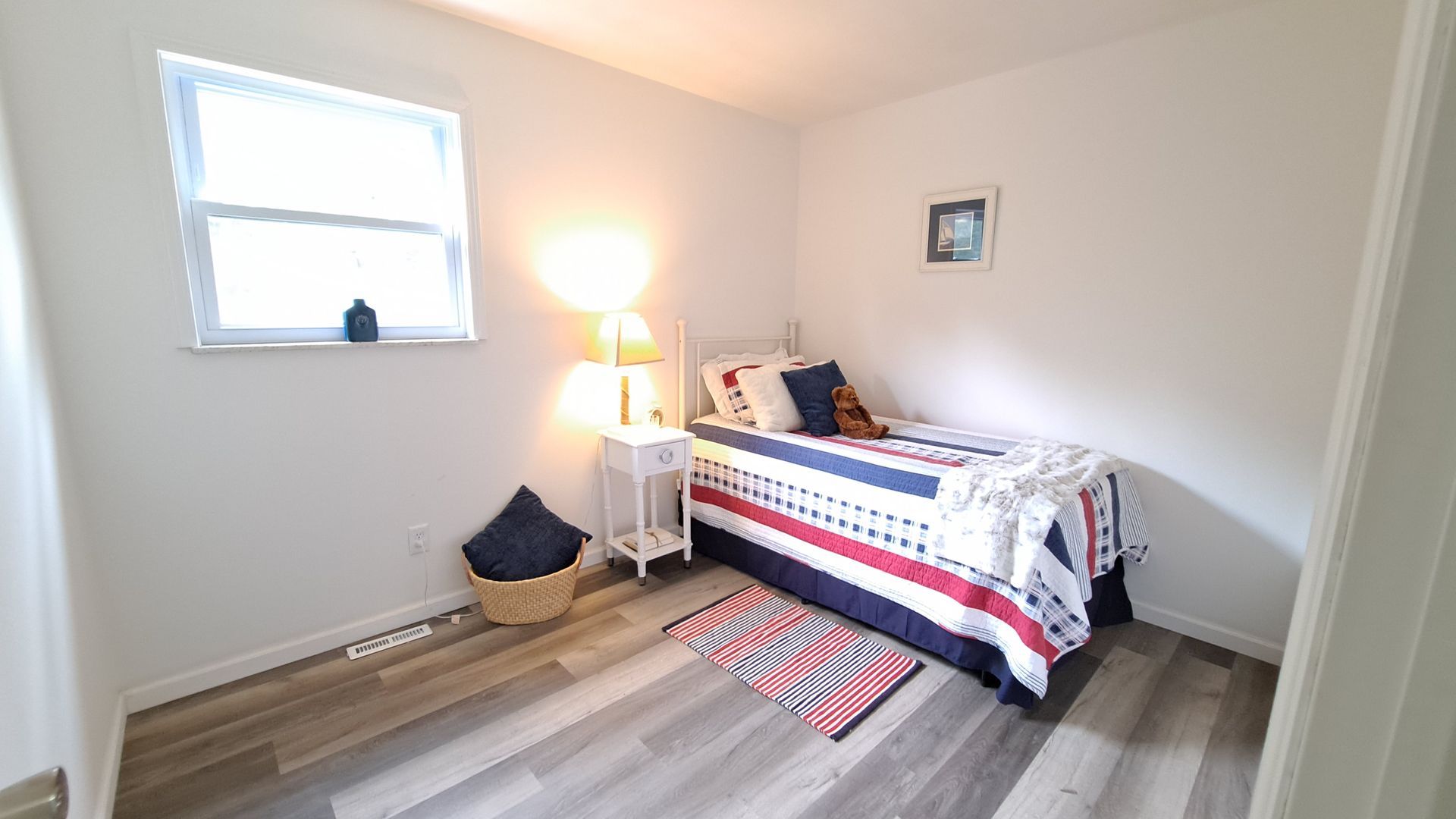 A small, white bedroom with a single bed and patriotic bedding. Window on the left, bedside table and rug.