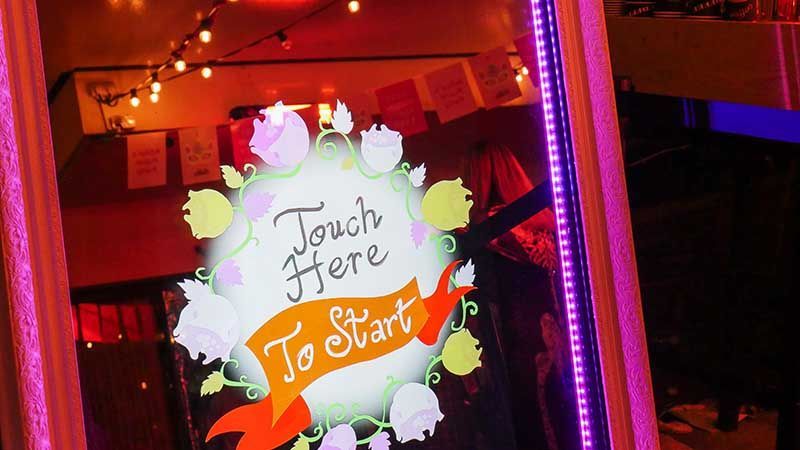 A photo booth mirror with "Touch Here To Start" graphic, surrounded by pink lights and floral decorations.