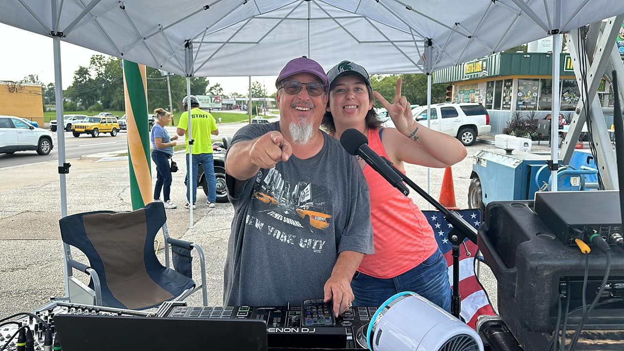Two people smile and point at the camera from a DJ setup under a tent.
