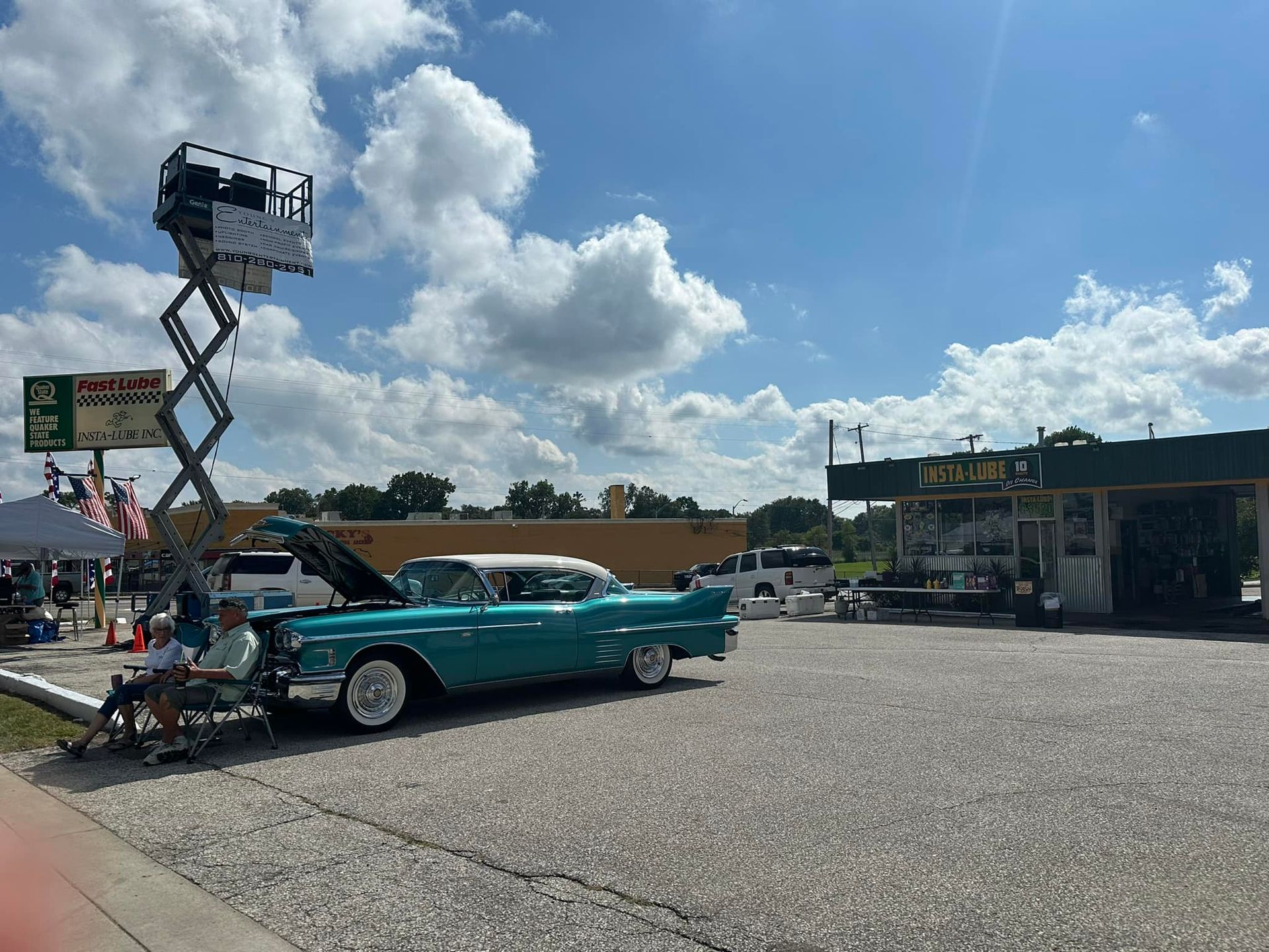 Teal classic car with hood open, at car show in front of a car wash, under blue sky.