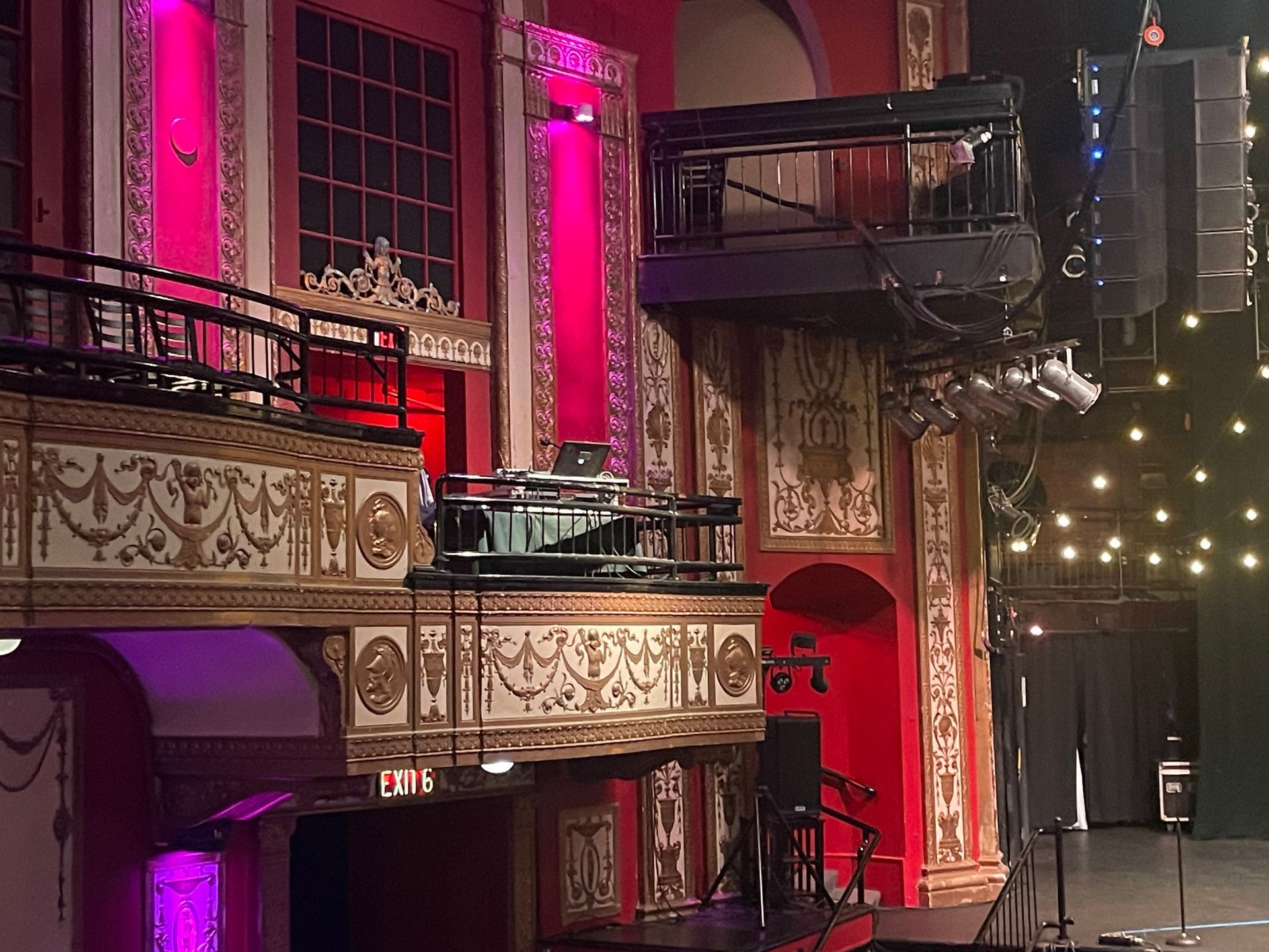 Theater interior with a balcony, stage, and sound equipment. Pink lighting and decorative gold accents.