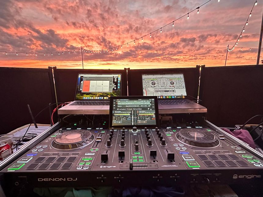 DJ setup outdoors at sunset, with two laptops and a tablet in front of a vibrant sky.