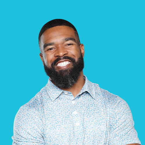 Man with a full beard smiling broadly, wearing a light blue patterned shirt against a blue background.