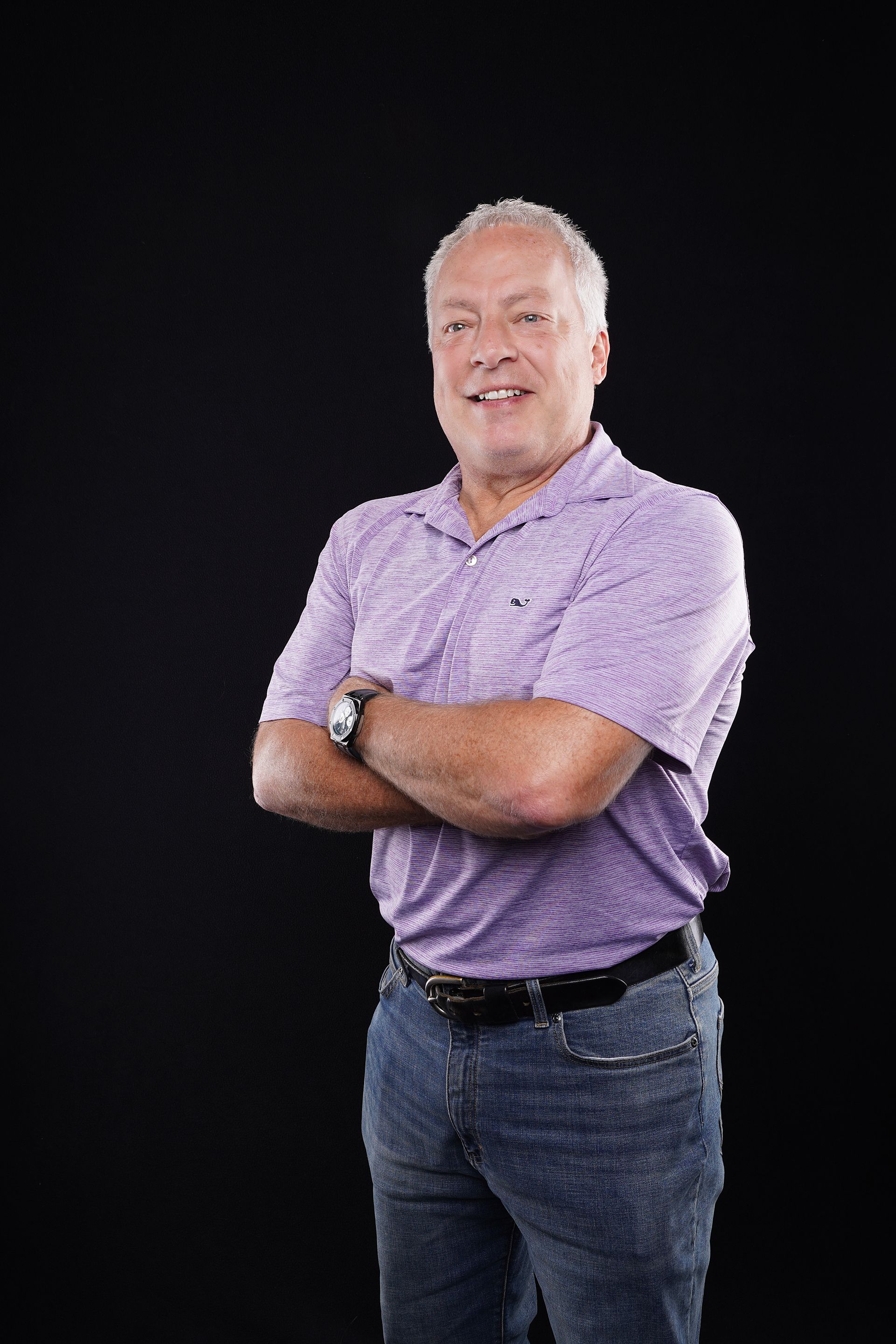 Man with silver hair, crossed arms, wearing purple shirt and jeans, smiling against black background.