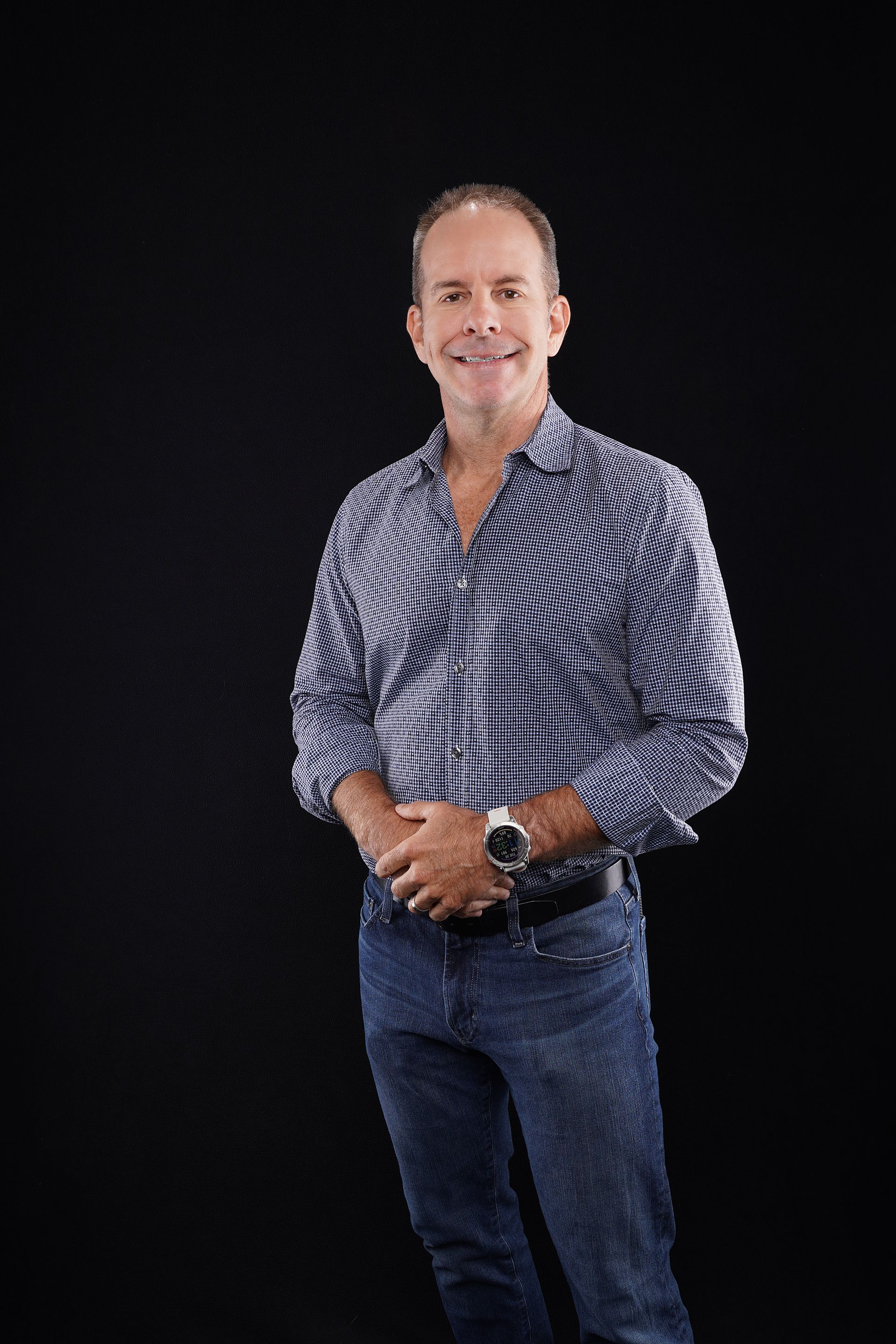 Man in patterned shirt and jeans, hands clasped, smiling against a black background.