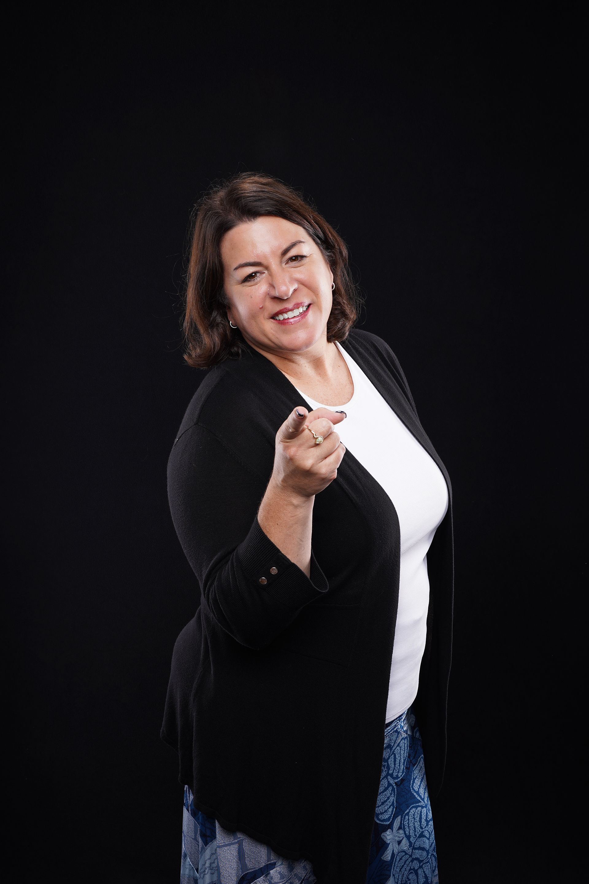 Woman pointing forward, smiling; black cardigan, white shirt, patterned skirt against black background.