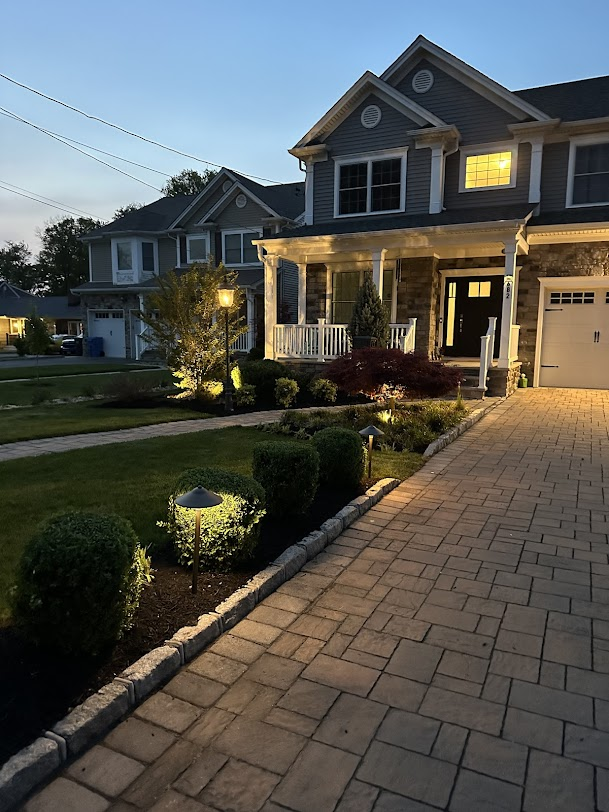 Two-story house with a brick driveway and illuminated landscaping at dusk.