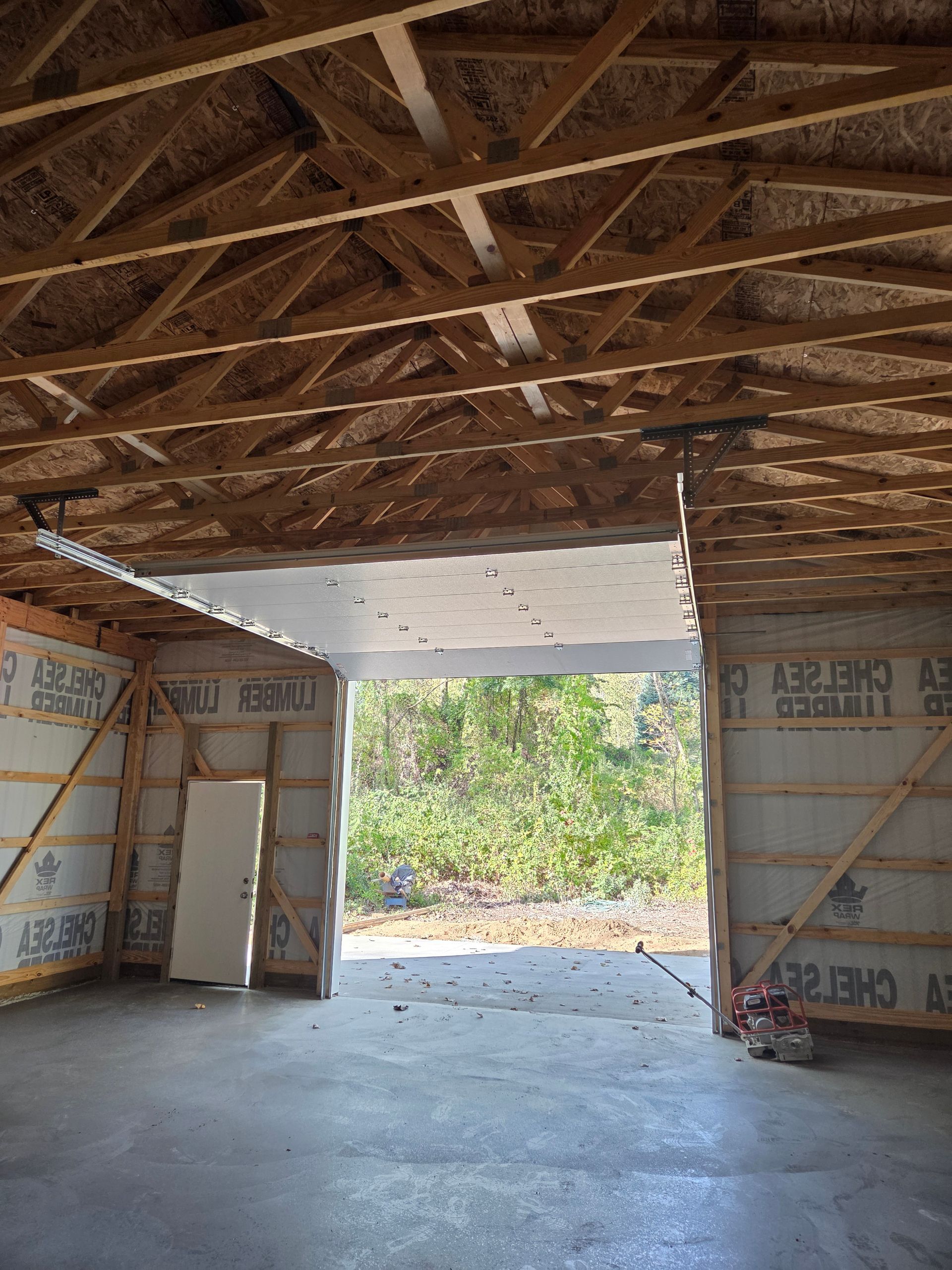 Interior of an unfinished building with an open garage door entrance, wooden beams and insulated walls.