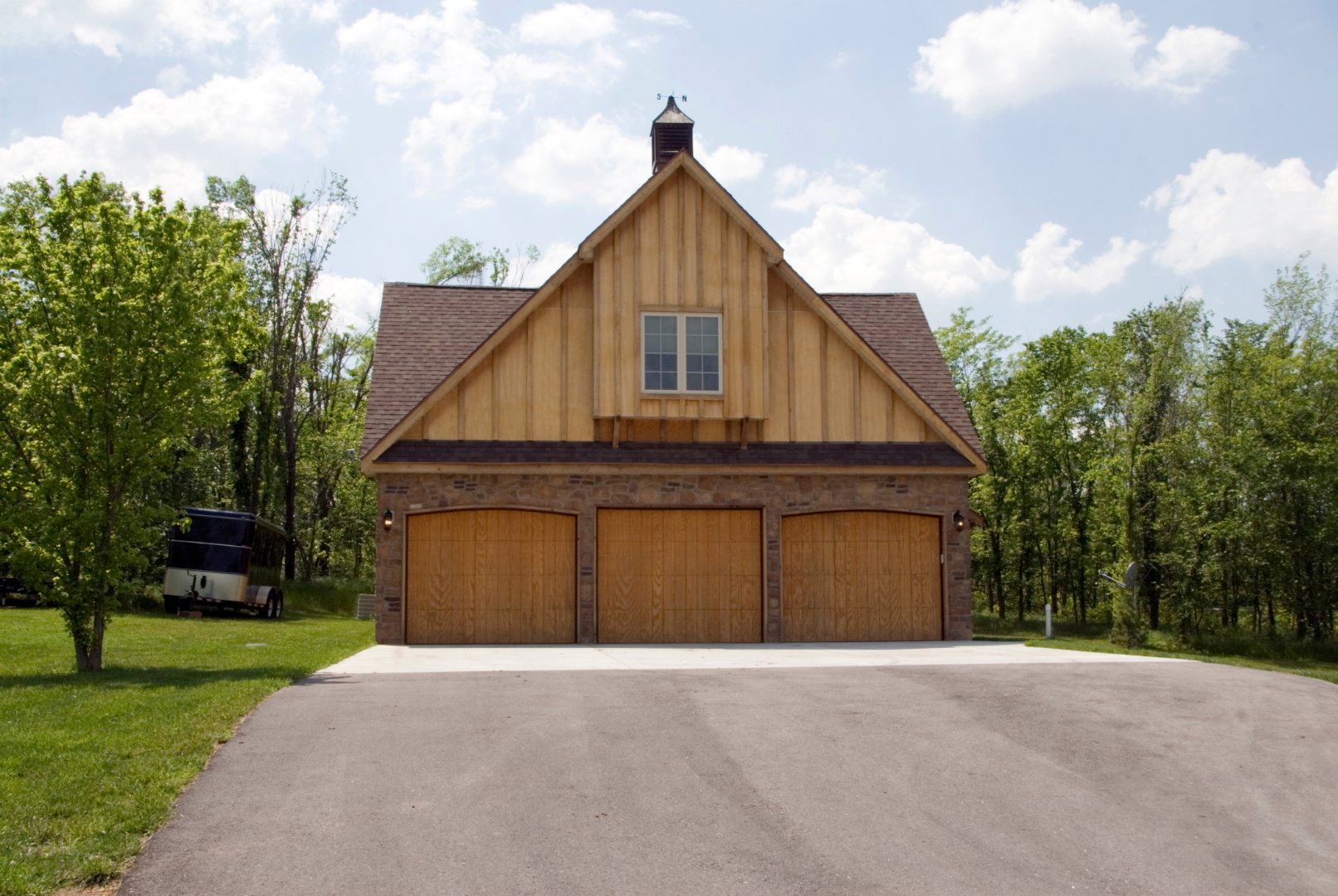 Three-bay garage with a barn-like facade, light brown wood, asphalt driveway, and trees on a sunny day.