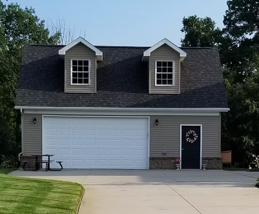 Two-story gray garage with white garage door, two dormers, and a dark door with a wreath.