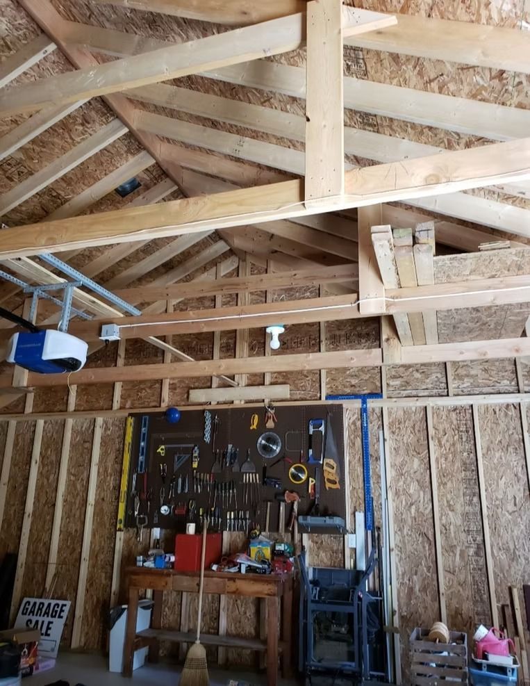 Interior of a garage with exposed wooden framing. Tools are hung on a pegboard. A garage door opener hangs on the ceiling.