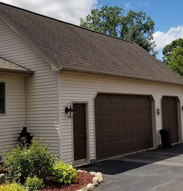 Beige house with brown roof, garage doors, and trim. Bushes and a trash can are present.