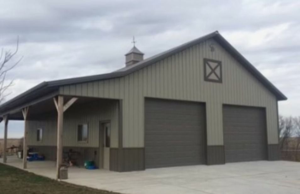 Tan and brown barn with a porch, two garage doors, and a cupola under a cloudy sky.