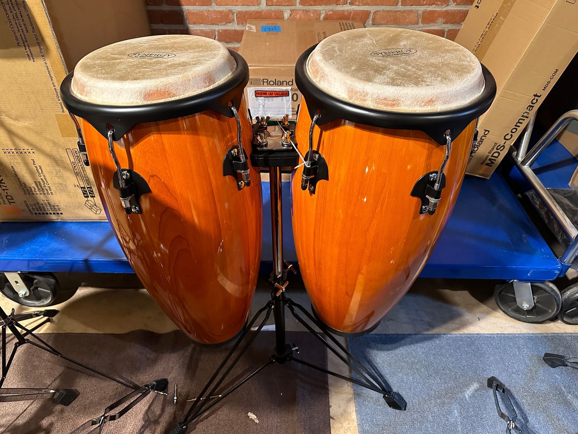 Two orange conga drums on a black stand, set against a cluttered background.