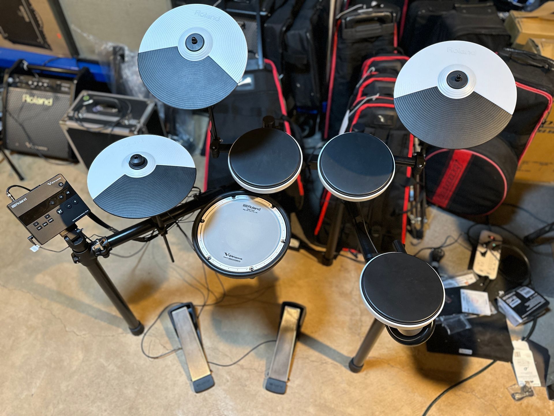 Electronic drum set with black and white pads and cymbals, pedals, module, and black cases in the background.