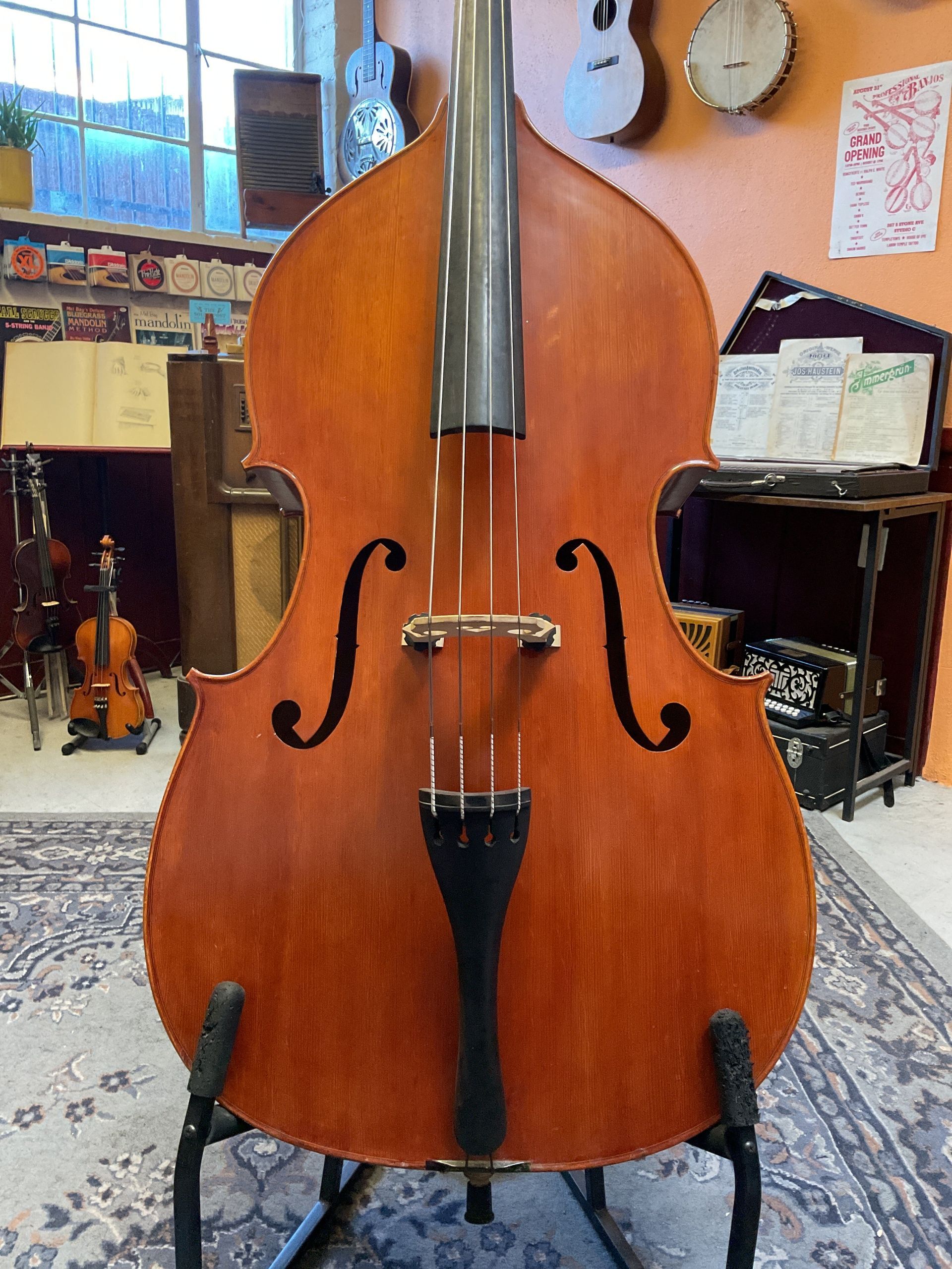 Upright bass on a stand in a room, with other instruments visible. Wooden instrument with black strings and bridge.